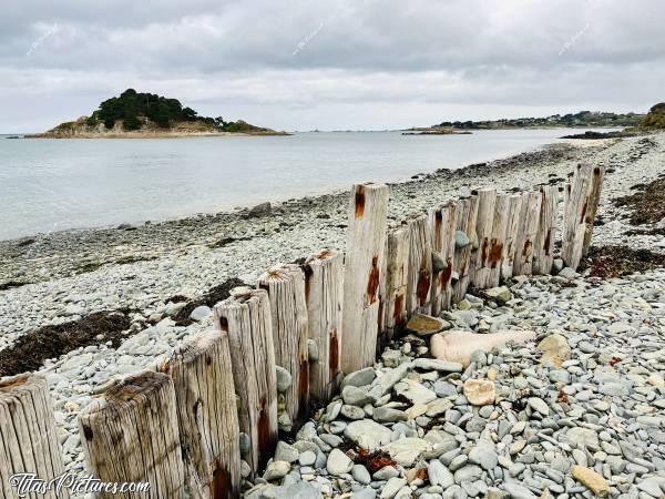 Photo Plouezoc’h : Petite randonnée pluvieuse à Plouezoc’h, dans la Baie de Morlaix. 
On ne le voit pas très bien sur la photo, en raison de la grisaille, mais les galets sont bleus clairs. C’est pas très courant ça 😅
Ces vieux poteaux de chemin de fer ont été dressés à cet endroit, pour protéger des vagues, l’escalier qui se trouve derrière moi. 
En face, l’Île Stérec.c, Plouezoc’h, plage, galets